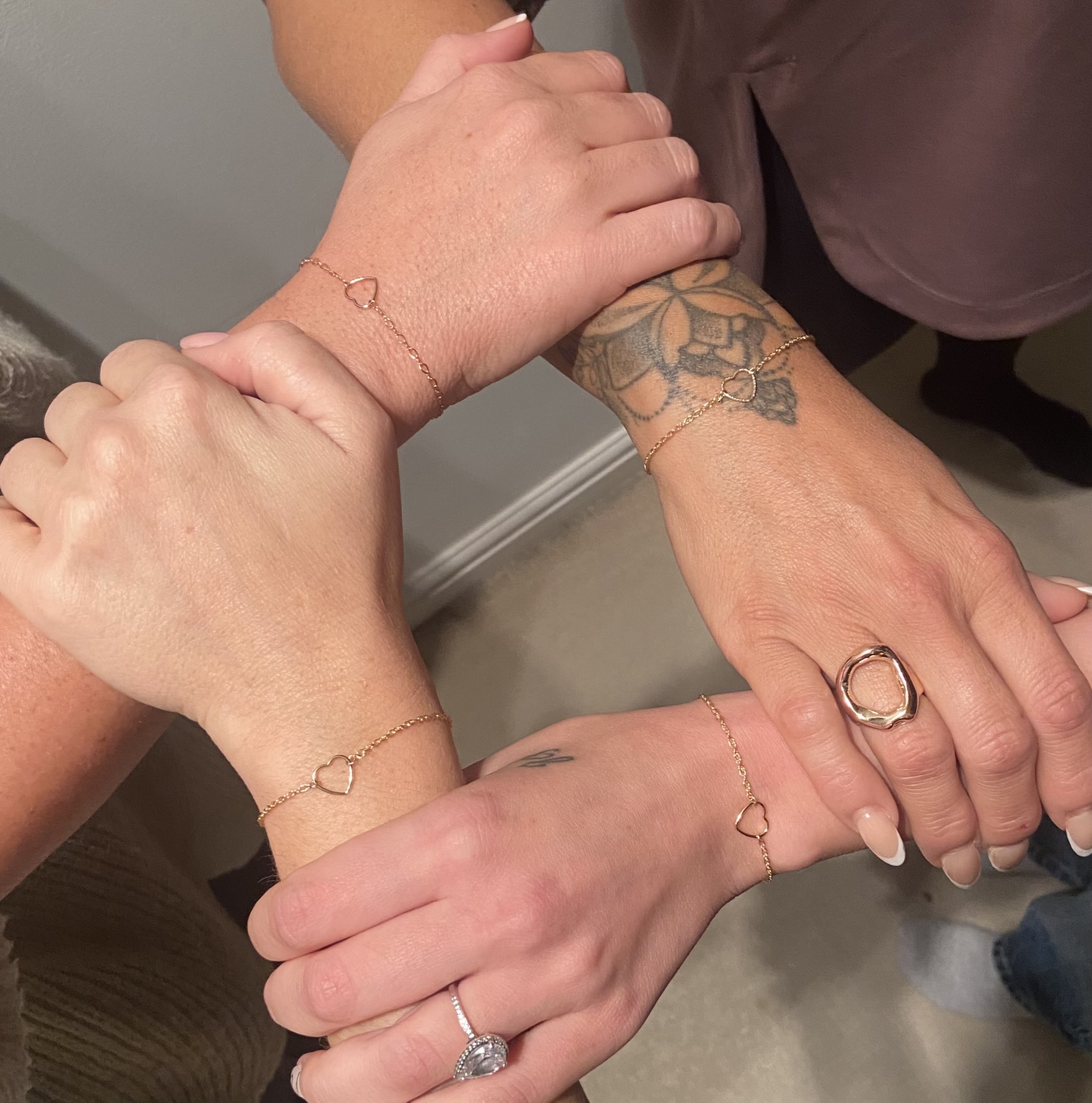 Four women clasp hands to show their matching heart charm permanent bracelets, custom-fitted during a private at-home jewellery experience i- celebrating lifelong friendship with personal style and shared meaning.
